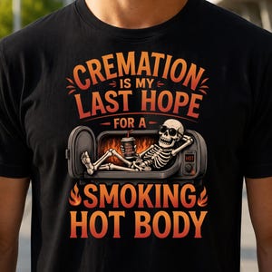 Op de afbeelding: Een zwart t-shirt met een afbeelding van een skelet met een zonnebril, liggend in een open oven terwijl het een drankje vasthoudt. De tekst luidt: CREMATION IS MY LAST HOPE FOR A SMOKING HOT BODY, met oranje en rode vlammen.