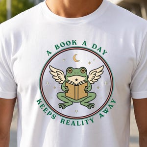 Peut inclure: Un t-shirt blanc avec un motif circulaire représentant une grenouille verte ailée lisant un livre ouvert. Le texte "A BOOK A DAY KEEPS REALITY AWAY" est imprimé en vert autour du cercle, qui contient aussi un croissant de lune et des étoiles.