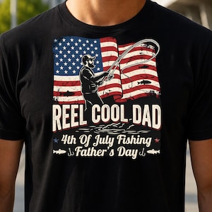 Peut inclure: T-shirt noir avec un motif représentant un drapeau américain et un pêcheur. Le texte indique "REEL COOL DAD" au-dessus de "4th Of July Fishing Father's Day". Le motif comprend des poissons et des hameçons.