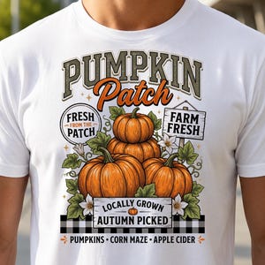 Peut inclure: Un t-shirt blanc avec une impression graphique de trois citrouilles orange, des feuilles vertes et des fleurs blanches. Le texte indique PUMPKIN PATCH, FRESH FROM THE PATCH, FARM FRESH, LOCALLY GROWN, AUTUMN PICKED, PUMPKINS, CORN MAZE et APPLE CIDER.