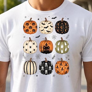 Peut inclure: Un t-shirt blanc avec une impression graphique de neuf citrouilles disposées en grille. Les citrouilles présentent divers motifs sur le thème d'Halloween, notamment des visages, des chauves-souris, des lunes, des étoiles, des araignées, une maison hantée, des fantômes, des rayures, des crânes et des chapeaux de sorcière.