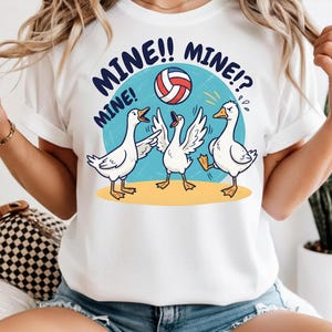 Peut inclure: T-shirt blanc avec une illustration de dessin animé de trois oies jouant au volley-ball. Les oies sont sur une surface jaune sur un fond bleu, avec les mots "MINE!! MINE!? MINE!" en bleu marine.