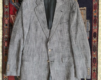 May include: A grey blazer with a textured weave, featuring a notched lapel, two front pockets, and three buttons on each cuff. The blazer has a two-button closure and a dark lining. The garment is laid flat on a patterned rug.