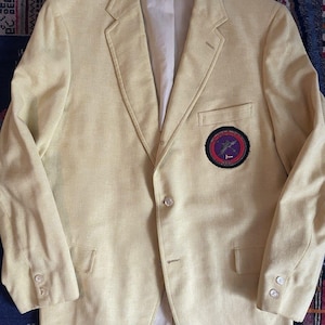 May include: A light yellow blazer with a single-breasted design. The blazer features a patch on the chest with a circular emblem in shades of purple, red, and black. It has two front pockets and buttoned cuffs.