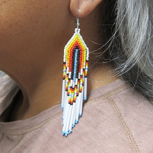 May include: A pair of long, beaded earrings with a white, red, yellow, blue, and black pattern. The earrings are hanging from a person's ear.