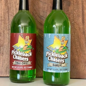 May include: Two green glass bottles with labels featuring a cartoon woman sitting on a pickle. The labels read "Jula's Pickleback Chasers" with the flavours "Thai Chili" and "Garlic". Each bottle contains 750 ml.