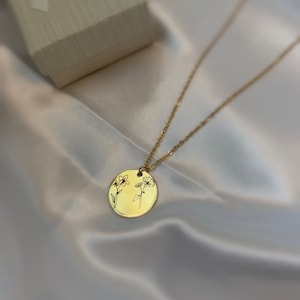 May include: A gold-colored necklace with a circular pendant featuring etched floral designs. The pendant hangs from a delicate chain, and a small gift box is visible in the background. The necklace rests on a white satin surface.