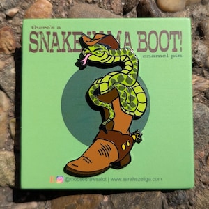 Op de afbeelding: Groene slang die een bruine cowboylaars met sporen draagt. De slang draagt een bruine cowboyhoed. De tekst "there's a SNAKE IN MY BOOT! enamel pin" staat op de groene achtergrond.