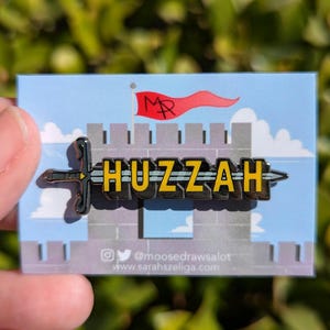 Puede incluir: Un pin de esmalte negro y amarillo que representa una espada con la palabra "HUZZAH" en ella. La espada está delante de un castillo de dibujos animados con una bandera roja que dice "MR".