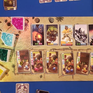 Peut inclure: Un plateau de jeu avec des gemmes color&eacute;es, des cartes et des illustrations. Le plateau est en bois et a une couleur brun clair. Les gemmes sont de diff&eacute;rentes couleurs, notamment rose, bleu, vert et jaune. Les cartes sont &eacute;galement de diff&eacute;rentes couleurs et pr&eacute;sentent des illustrations de personnes, d'animaux et d'objets. Le plateau est configur&eacute; pour un jeu de gestion des ressources et d'&eacute;change.