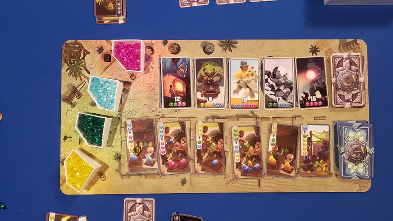 Peut inclure: Un plateau de jeu avec des gemmes color&eacute;es, des cartes et des illustrations. Le plateau est en bois et a une couleur brun clair. Les gemmes sont de diff&eacute;rentes couleurs, notamment rose, bleu, vert et jaune. Les cartes sont &eacute;galement de diff&eacute;rentes couleurs et pr&eacute;sentent des illustrations de personnes, d'animaux et d'objets. Le plateau est configur&eacute; pour un jeu de gestion des ressources et d'&eacute;change.