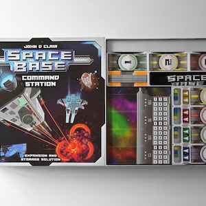 May include: An open board game box set, "Space Base: Command Station", with a space theme. The box displays game components, including cards, tokens, and a game board. The box art features spaceships and a cosmic background.