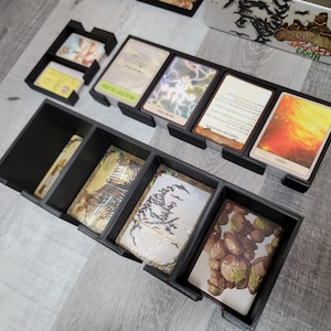 May include: Black plastic organiser with compartments for storing cards. The organiser has a variety of compartments of different sizes. The compartments are filled with cards with images of fantasy creatures and landscapes.