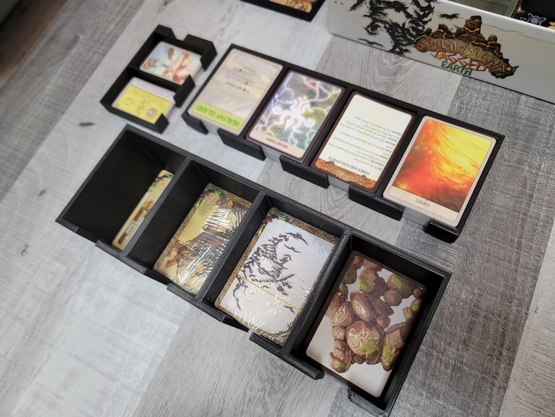 May include: Black plastic organiser with compartments for storing cards. The organiser has a variety of compartments of different sizes. The compartments are filled with cards with images of fantasy creatures and landscapes.