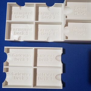 May include: White 3D-printed game card organizers. The organizers are labeled with text such as "Farmyard Deck 1", "Farmyard Deck 2", "Point Deck", "Bonus Deck", "Gateway Deck 1", and "Gateway Deck 2". The organizers are designed to hold game cards.