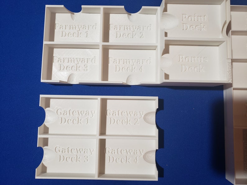 May include: White 3D-printed game card organizers. The organizers are labeled with text such as "Farmyard Deck 1", "Farmyard Deck 2", "Point Deck", "Bonus Deck", "Gateway Deck 1", and "Gateway Deck 2". The organizers are designed to hold game cards.