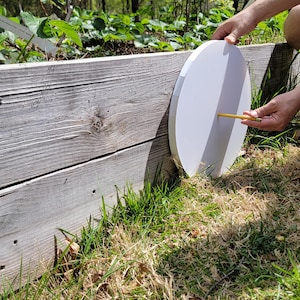 Op de afbeelding: Een persoon gebruikt een potlood om een wit rond object te markeren dat tegen een houten plantenbak is geplaatst. Het object bevindt zich gedeeltelijk in de schaduw en gedeeltelijk in de zon.