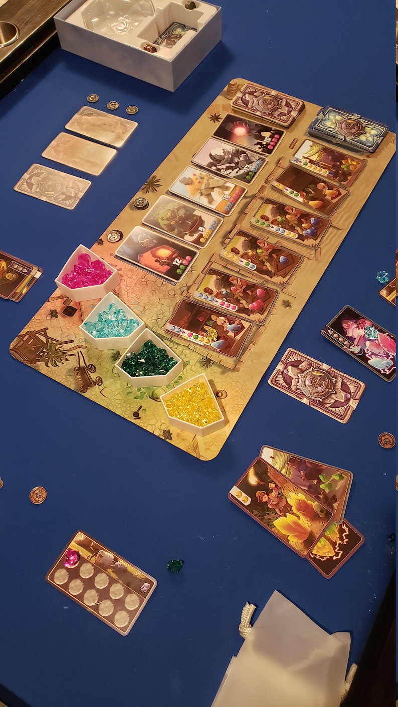 Peut inclure: Un plateau de jeu avec des gemmes color&eacute;es et des cartes. Le plateau est en bois et a un sch&eacute;ma de couleurs brun et beige. Les cartes sont de couleurs diff&eacute;rentes et ont des images de personnes et d'animaux dessus. Il y a aussi de petits contenants blancs avec des gemmes &agrave; l'int&eacute;rieur.