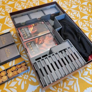 May include: A board game organizer with compartments for cards, tokens, and other game components. The organizer is made of gray plastic and includes card dividers, card boxes, and a black drawstring bag. The game cards feature fantasy art.