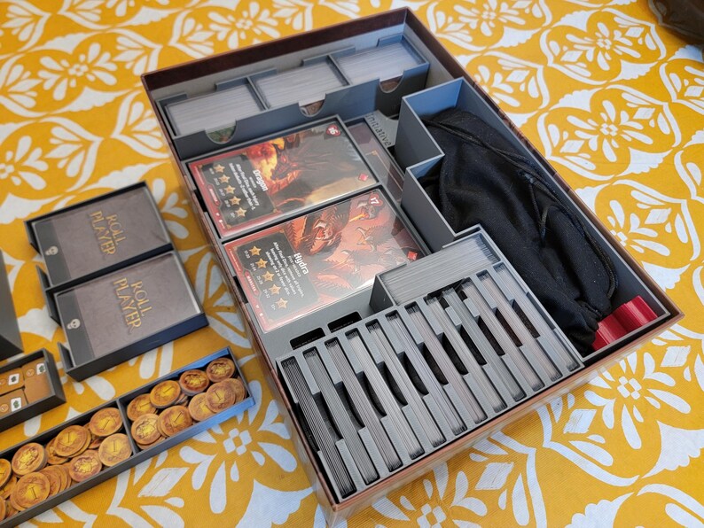 May include: A board game organizer with compartments for cards, tokens, and other game components. The organizer is made of gray plastic and includes card dividers, card boxes, and a black drawstring bag. The game cards feature fantasy art.