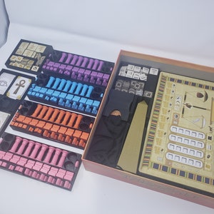 May include: A board game with a beige and brown game board, a black storage box, and multiple trays of colorful game pieces. The game board features a scale, a boat, and a hieroglyphic design. The trays contain pink, blue, orange, and purple game pieces.