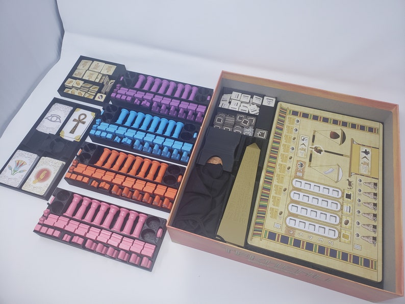 May include: A board game with a beige and brown game board, a black storage box, and multiple trays of colorful game pieces. The game board features a scale, a boat, and a hieroglyphic design. The trays contain pink, blue, orange, and purple game pieces.