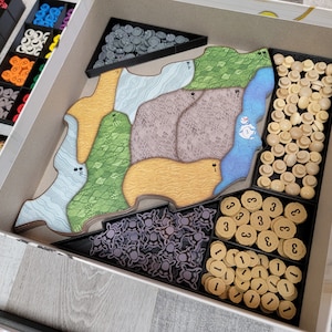 May include: A white box containing a variety of game pieces, including wooden tokens, plastic figures, and cardboard tiles. The box is divided into compartments, each containing a different type of piece. The pieces are arranged neatly and in an organised manner.