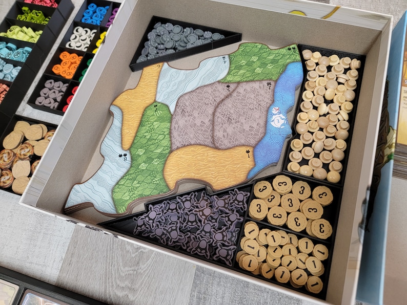 May include: A white box containing a variety of game pieces, including wooden tokens, plastic figures, and cardboard tiles. The box is divided into compartments, each containing a different type of piece. The pieces are arranged neatly and in an organised manner.