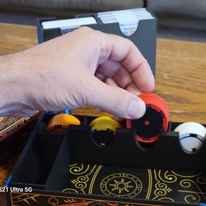 May include: A black organizer box with compartments for storing game pieces. The box has a gold and black design with a sun symbol in the center. There are red, yellow, and white game pieces in the compartments.