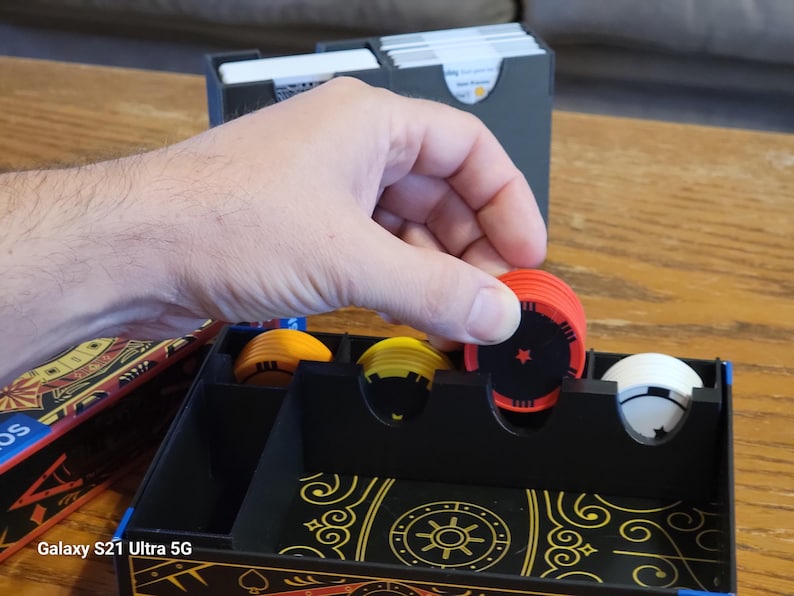 May include: A black organizer box with compartments for storing game pieces. The box has a gold and black design with a sun symbol in the center. There are red, yellow, and white game pieces in the compartments.