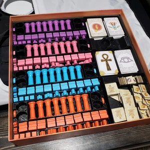 May include: A game box with a variety of colorful game pieces, including pink, purple, blue, and orange. The box also contains cards with images of an ankh, an eye, and a lotus flower.