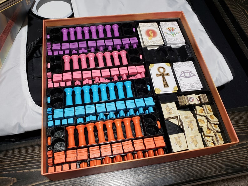 May include: A game box with a variety of colorful game pieces, including pink, purple, blue, and orange. The box also contains cards with images of an ankh, an eye, and a lotus flower.