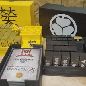 Puede incluir: Un juego de mesa con componentes amarillos y negros. Las piezas amarillas tienen caracteres japoneses. Las piezas negras tienen símbolos blancos. El juego incluye cartas con texto japonés y un emblema dorado. El juego está sobre un mapa.