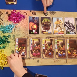 Peut inclure: Un plateau de jeu avec des gemmes color&eacute;es, des cartes et un jeton en bois. Les cartes pr&eacute;sentent des illustrations de personnes, d'animaux et d'objets. Le plateau est en mat&eacute;riau brun clair et a une texture de sable.