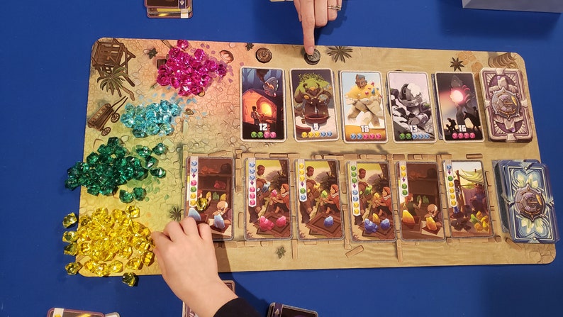 Peut inclure: Un plateau de jeu avec des gemmes color&eacute;es, des cartes et un jeton en bois. Les cartes pr&eacute;sentent des illustrations de personnes, d'animaux et d'objets. Le plateau est en mat&eacute;riau brun clair et a une texture de sable.