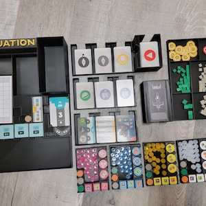 May include: A black organizer with the word "EVACUATION" printed on the front. The organizer has compartments for storing game pieces, cards, and other accessories. The organizer is filled with various colored game pieces, including yellow, red, green, and blue.