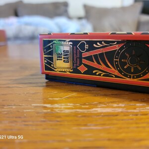 May include: A red, black, and gold box with the text "The Gang" and "The Cooperative Poker Game" on the front. The box is on a wooden surface.
