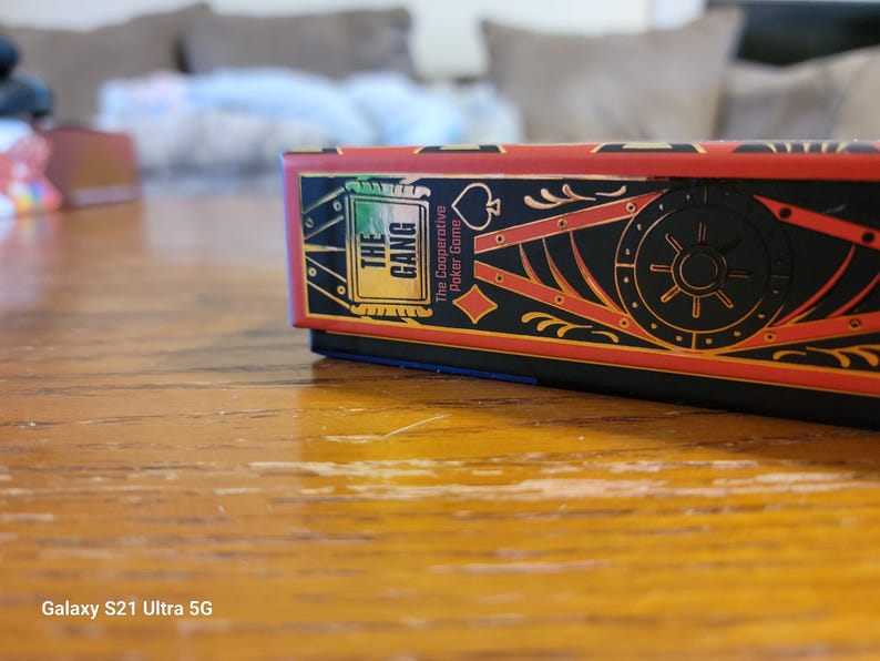May include: A red, black, and gold box with the text "The Gang" and "The Cooperative Poker Game" on the front. The box is on a wooden surface.