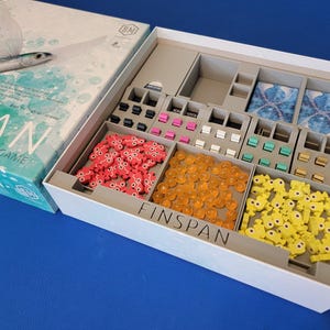 Könnte beinhalten: Eine weiße Box mit der Aufschrift "Finspan" auf der Vorderseite. Die Box ist geöffnet und enthält verschiedene Spielsteine, darunter kleine rote, gelbe und blaue Plastikteile sowie eine Reihe orangefarbener Glasperlen. Die Box ist Teil eines Brettspiels namens "Wingspan".