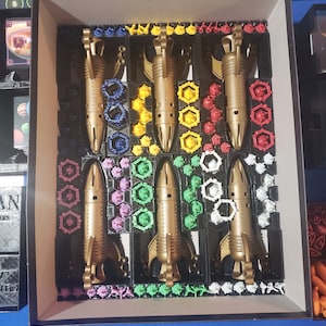 May include: A Catan board game insert with five gold spaceships, each with a different colored base. The spaceships are surrounded by small colored plastic pieces.
