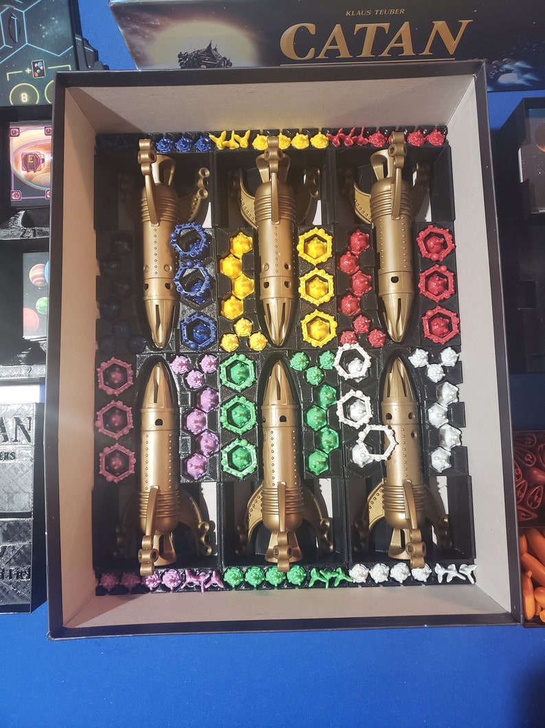 May include: A Catan board game insert with five gold spaceships, each with a different colored base. The spaceships are surrounded by small colored plastic pieces.