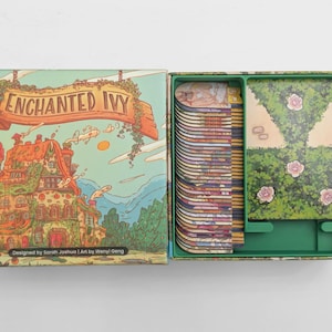 May include: A board game box titled Enchanted Ivy with a colorful illustration of a house covered in vines. The open box contains a stack of vertical game cards and a larger rectangular game board featuring a green hedge path with pink roses.