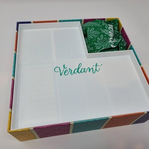 Puede incluir: Una caja blanca con un patr&oacute;n de rayas de colores en el exterior. La caja tiene una tapa extra&iacute;ble y un compartimento en el interior. La caja tiene la palabra "Verdant" escrita en el interior.