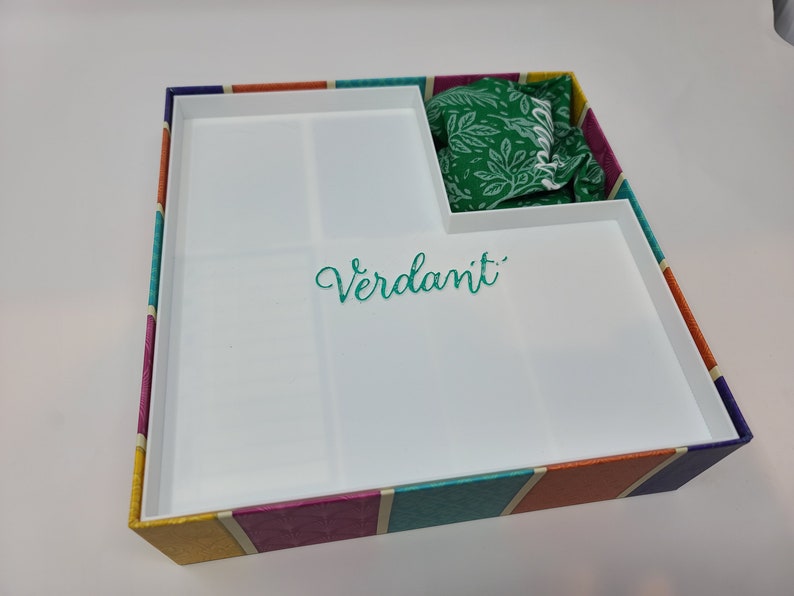 Puede incluir: Una caja blanca con un patr&oacute;n de rayas de colores en el exterior. La caja tiene una tapa extra&iacute;ble y un compartimento en el interior. La caja tiene la palabra "Verdant" escrita en el interior.