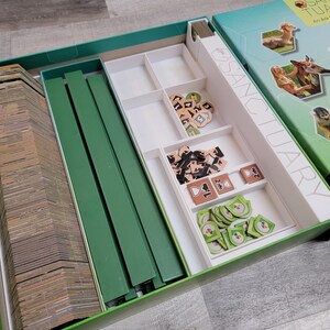 May include: Open board game box with the title "Sanctuary" visible. The box is teal and green, with images of animals on the lid. Inside, there are stacks of cards, green dividers, and various game pieces in white compartments.