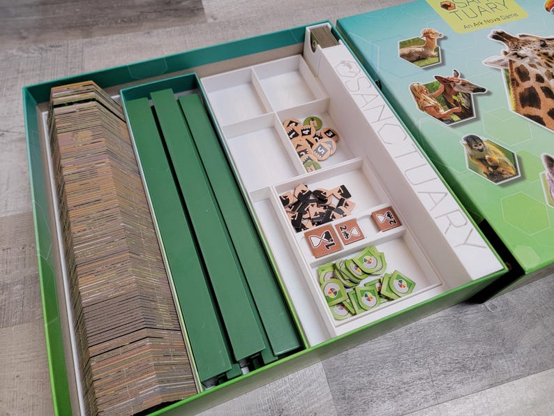 May include: Open board game box with the title "Sanctuary" visible. The box is teal and green, with images of animals on the lid. Inside, there are stacks of cards, green dividers, and various game pieces in white compartments.