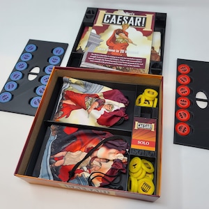 Puede incluir: Una caja de juego con un diseño rojo y dorado. La caja está abierta y muestra el contenido, incluyendo dos bolsas de tela, una tarjeta que dice "¡César! Solo", y un conjunto de fichas amarillas. La caja también tiene un inserto negro con compartimentos para las piezas del juego.