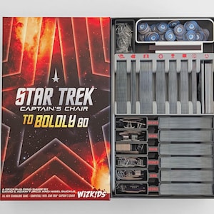 May include: Star Trek Captain's Chair To Boldly Go board game box with a red and orange space-themed cover featuring a large star graphic. The open box reveals a gray plastic insert containing organized stacks of cards and blue circular tokens.