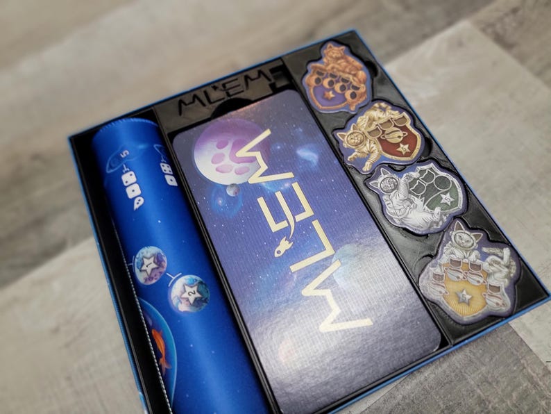 Peut inclure: Une bo&icirc;te de jeu de soci&eacute;t&eacute; bleue et dor&eacute;e avec le titre "Meow Meow" sur le couvercle. La bo&icirc;te contient un plateau de jeu bleu, un gobelet &agrave; d&eacute;s bleu et cinq pions sur le th&egrave;me des chats.