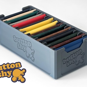 May include: A gray rectangular storage box with multiple fabric swatches in various colors. The box has the text "button shy" on the side and a logo with the same text and a yellow and orange design.